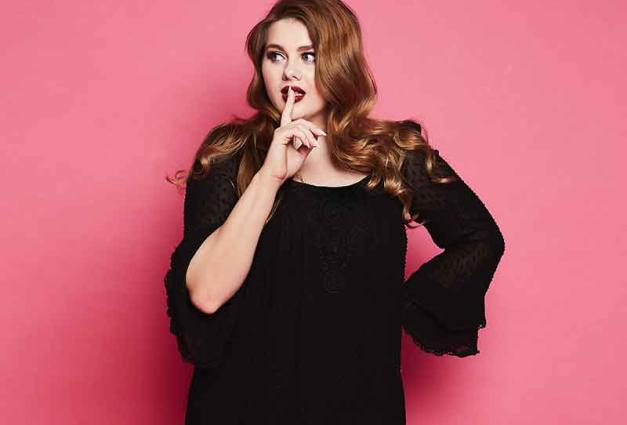 Plus size model in black short dress saying hush and posing at pink background
