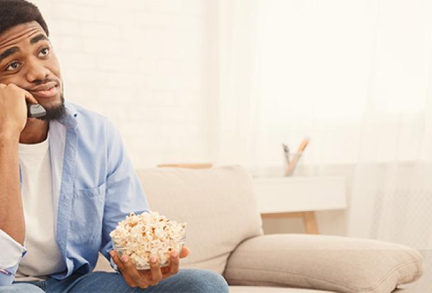 Man sitting on soaf holding remote control and bowl of popcorn looking indecisive about what to watch