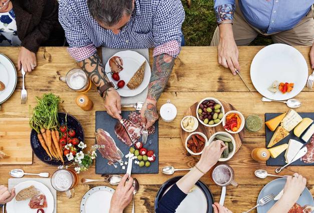 group of people eating otuside at a big table, some are men, some are women, young and old. boards of food and shared plates show people eating.