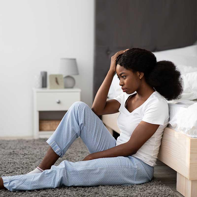Stressed young black woman sitting on floor by bed at home