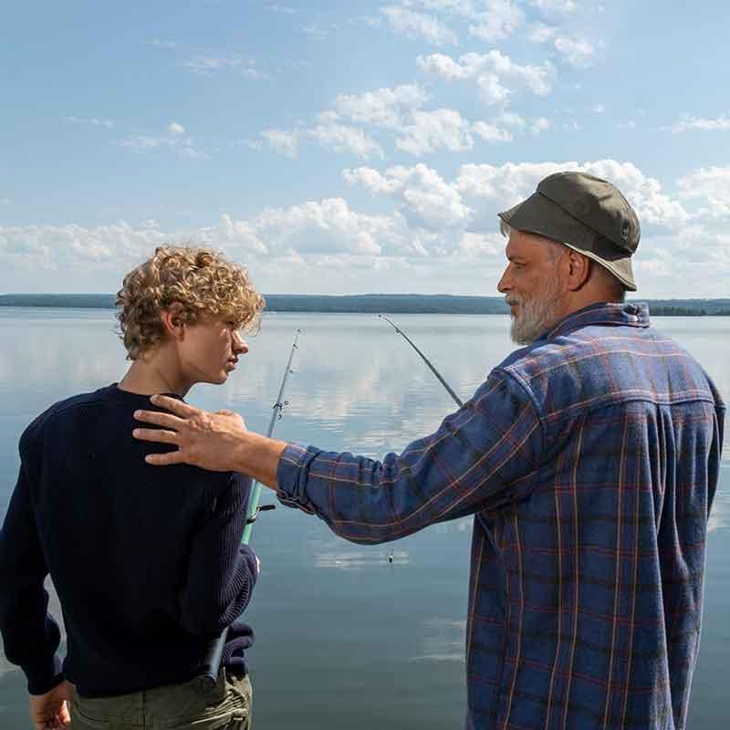 Older man touching teenage boy on shoulder near water while fishing