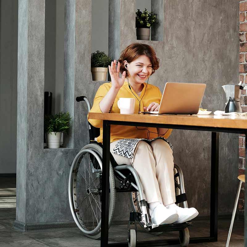 Woman in wheelchair at table waving and looking at a laptop computer